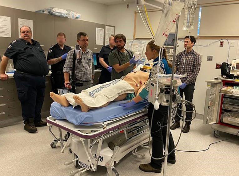 A group of paramedics and health care professionals stand in a medical room around a simulation mannequin.