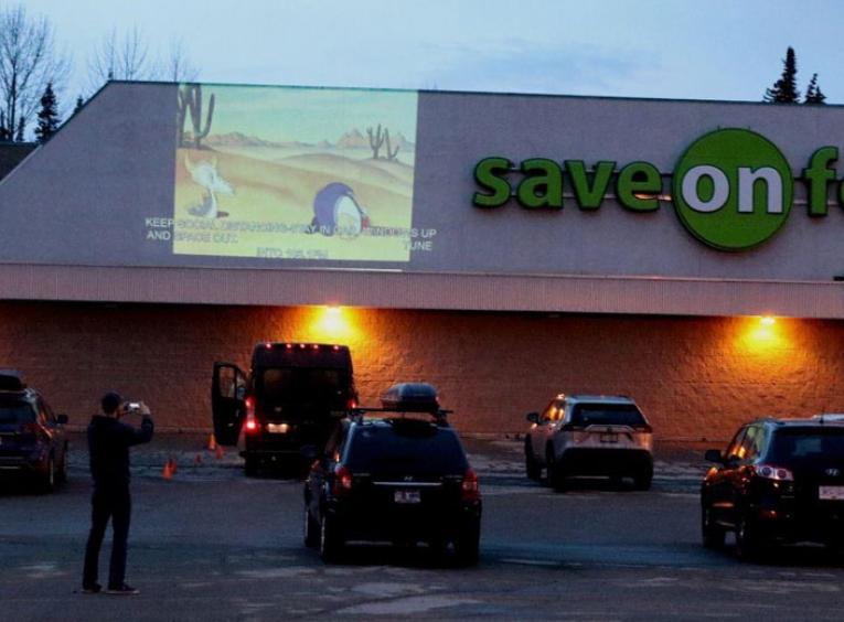 Cars are parked in a Save On Foods parking lot, a cartoon is projected on the wall.