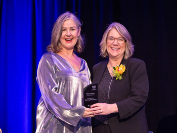 Two women stand smiling, while one presents the other with an award
