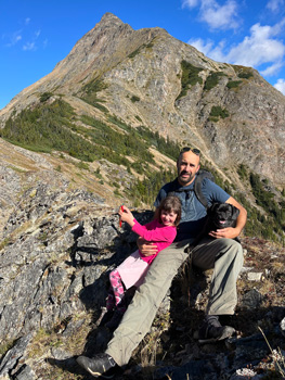 Man sitting with his daughter with a large mountain behind them.