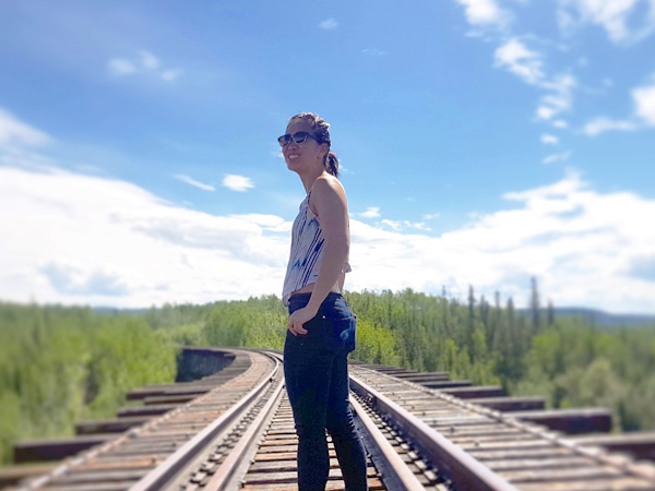 A woman stands on a train track that disappears in the distant forest. A sunny sky beats down on her.