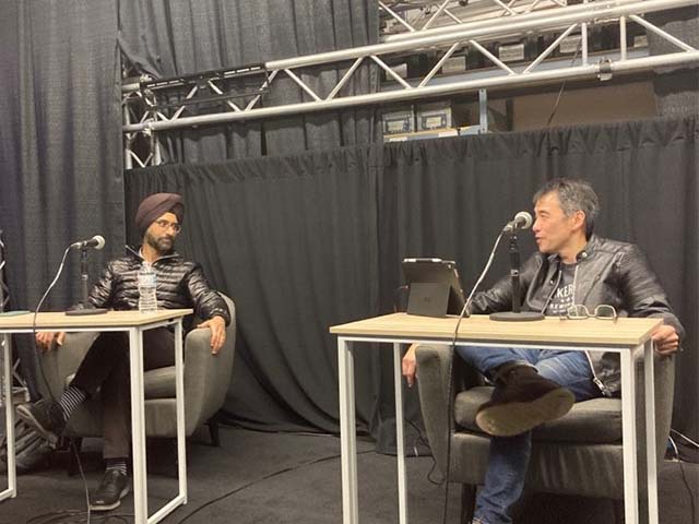 two men sit at a table with a black curtain behind them. they are recording a podcast episode