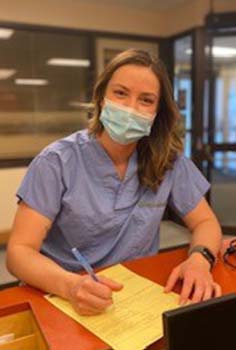 a woman with brown hair in scrubs and wearing a mask stands at a hospital desk
