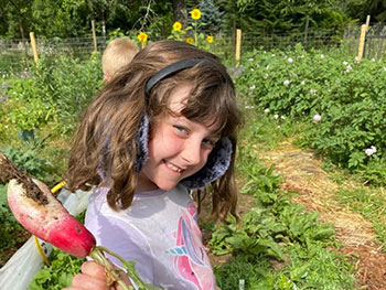 Child with long hair and wearing earmuffs holds up large radish