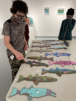 Youth stand looking at painted wooden fish laid out on a table to dry. 