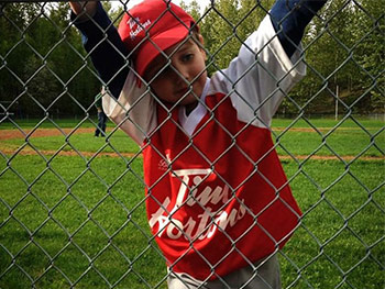 Little boy in baseball uniform leaning on a metal fence.