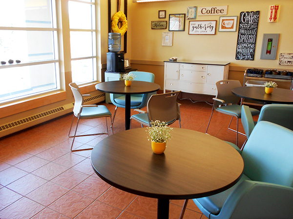 Several round tables with flowers on them, and chairs around them, are shown. The back wall has several coffee-themed signs hanging.