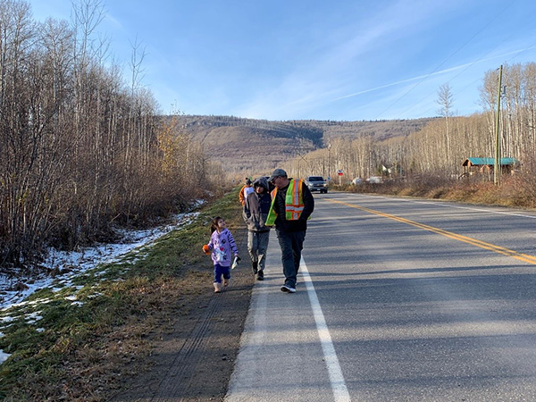 A man in a high vis vest walks along the side of a road with a young girl who is looking up at him.