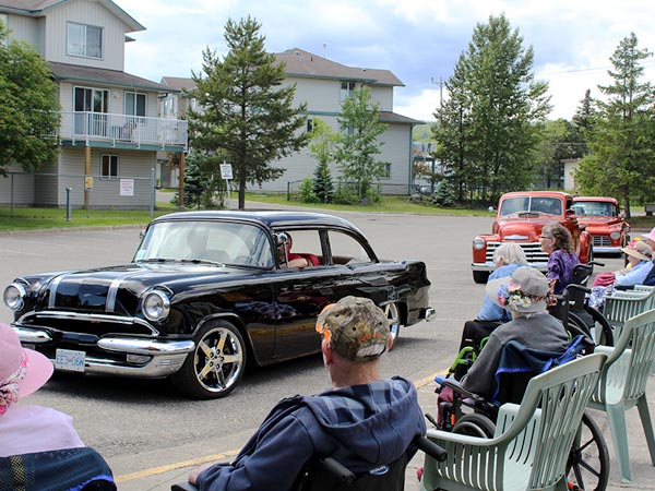 A group of seniors watch a line of classic cars drive by. 