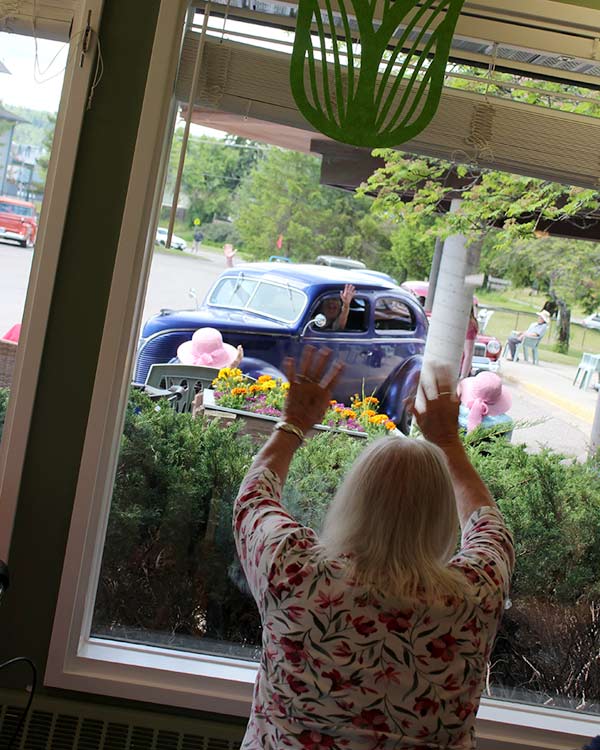 From inside a long-term care facility, a woman waves as classic cars drive through a parking lot. The driver of a blue car waves back. 