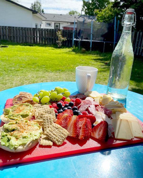 Avocado toast, fruits, meats, cheeses, and a bottle of water are on a cutting board on a backyard table. 