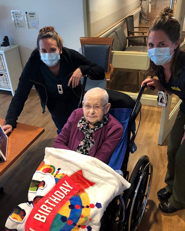 Two staff members wearing masks stand around an elderly woman in a wheel chair. 