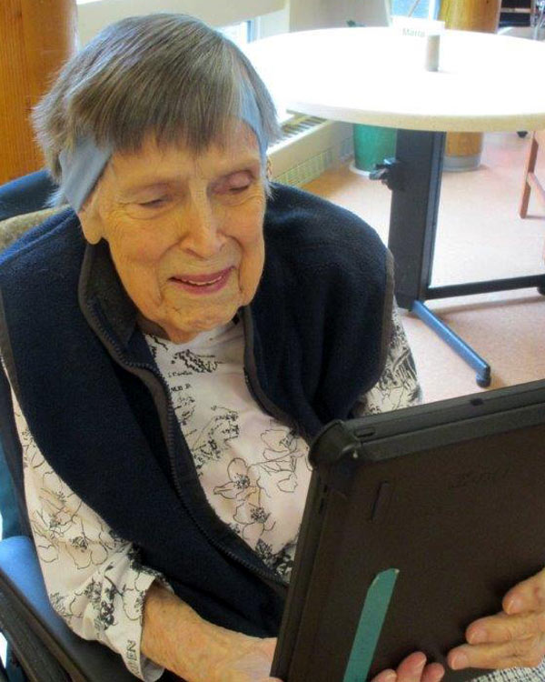 An elderly woman in a wheel chair smiles as she operates an iPad. 