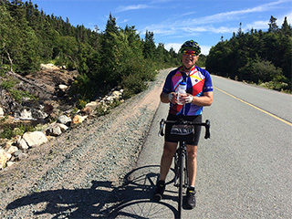Garry Knoll standing with his bicycle wearing a helmet and holding a water bottle on a road in the summer. 