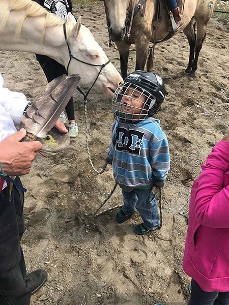 A young boy wearing a hockey helmet, holding a rope tied to a horse, looks up at a man holding feathers and items for smudging.