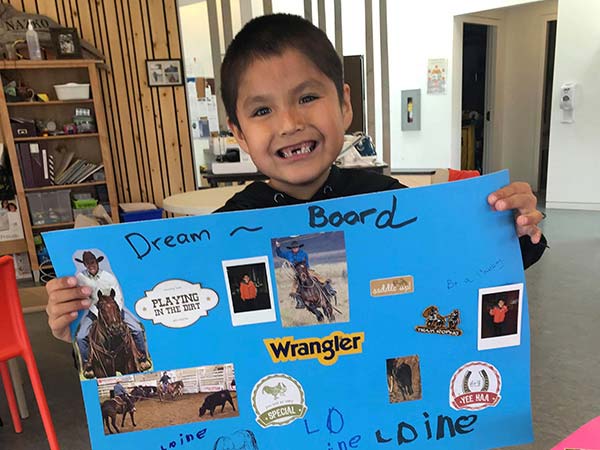 A young boy holds up a poster board, titled Dream Board, with magazine cut outs of horses and cowboys.