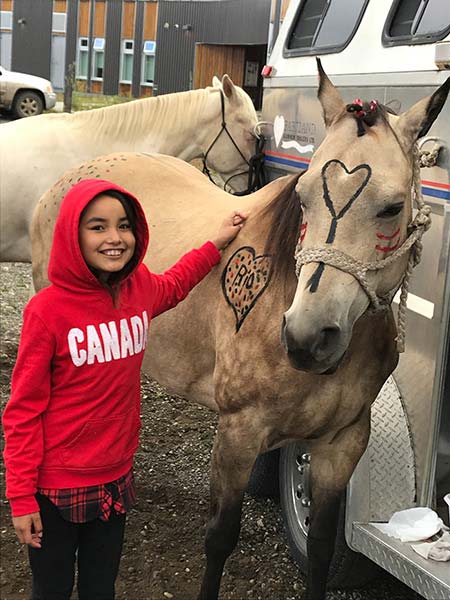 A young girl in a red hoodie stands next to a horse that is decorated with paint.