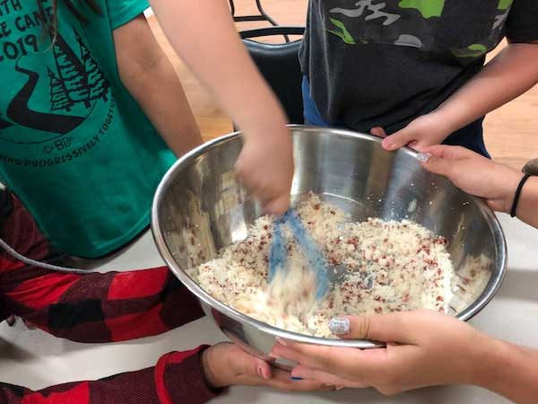Several campers hold a mixing bowl while another camper tosses the food with tongs.