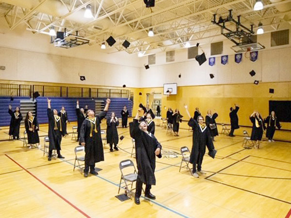 Standing a safe distance apart, students in a gym toss their grad caps into the air. 