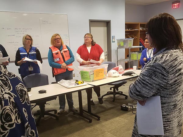 People stand around a meeting room table wearing vests and holding papers.