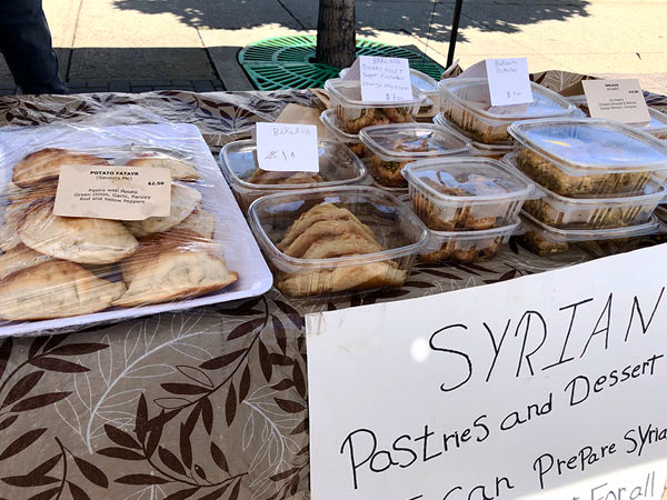 A variety of pastries and deserts are on a farmer's marker table. A sign says they are Syrian. 