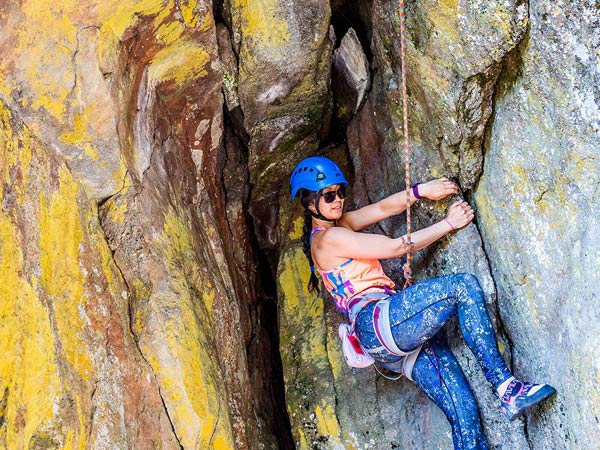 Melinda mountain climbs, suspended by a rope, hanging onto a rock. The rock fades from grey to brown and yellow.