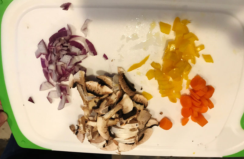 Red onions, mushrooms, yellow peppers, and carrots are shown on a cutting board. 