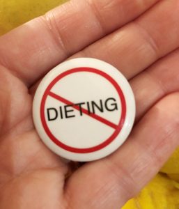 A hand holding a button that says Dieting with a line through it.