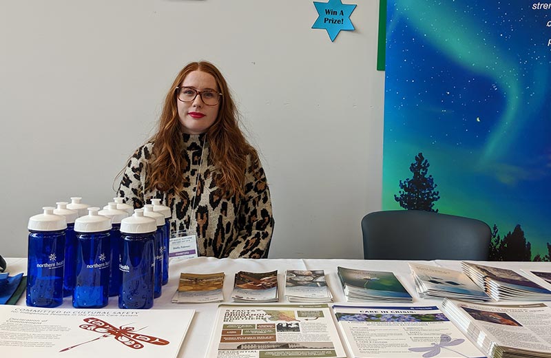 A woman sits behind a table with Indigenous Health pamphlets on it.