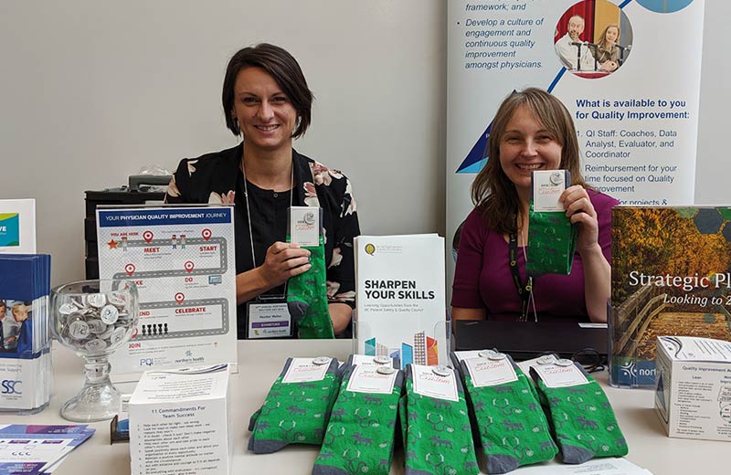 Two women sit behind a table that is covered in health care pamphlets.