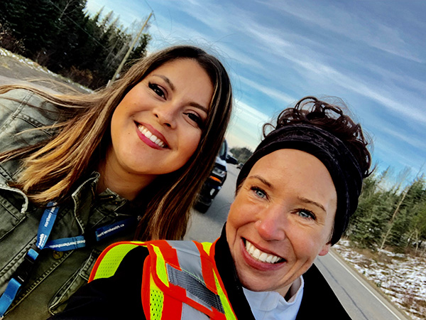 Two smiling women pose for a selfie outside on a road lined with snow and trees.