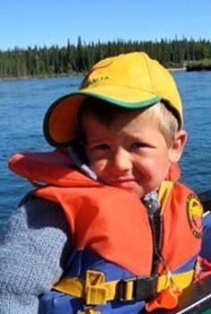 a small boy with blond hair sits in a boat with a red pfd on