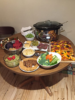 Potluck items on table including raw vegetables, guacamole, crackers, meat, cheese and olives.