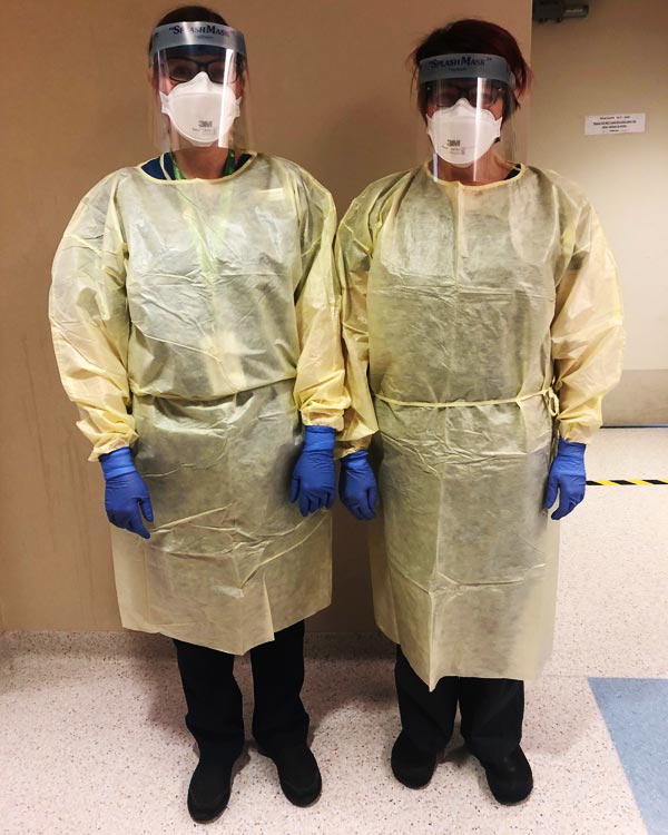 Two health care professionals wear gloves, gowns, and face masks with eye shields. 