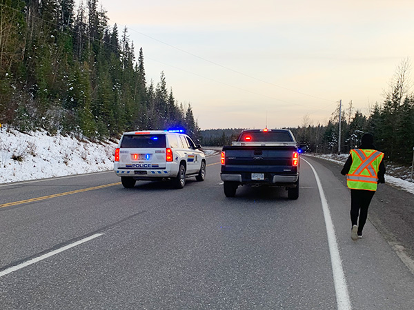 A police SUV with lights flashing and an unmarked police truck on the road, next to a walker.