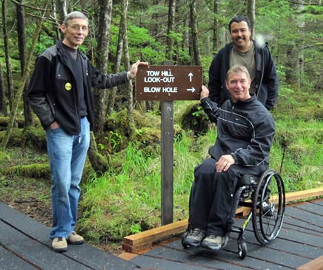 Two men standing next to another man in a wheelchair on an accessible forest park trail.