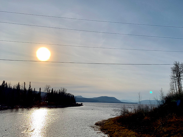 A view of a tree lined lake, hills on the horizon, and the sun shining in the sky.