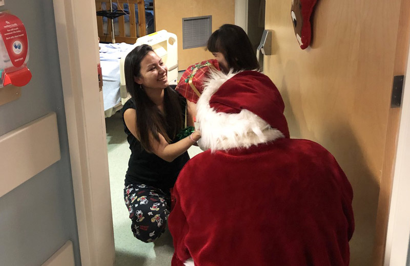 Santa crouched in front of a young girl with her mother, handing them a wrapped gift. 