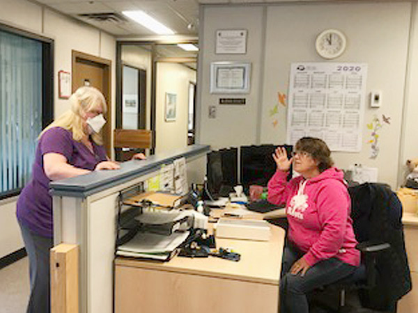 A woman wearing a medical mask and woman at a desk try to communicate. 