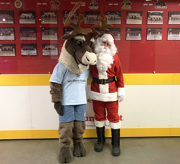 Man dress in a red and white Santa suit standing beside Spirit the Caribou mascot.