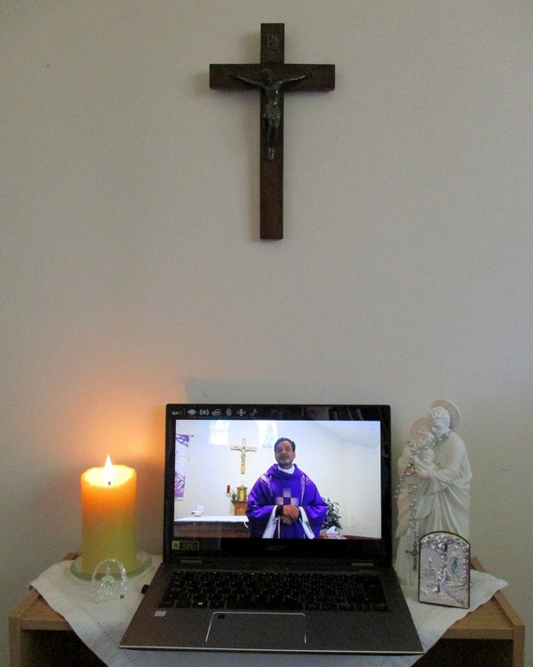 A church service streams on a laptop. A cross hangs on the wall above.