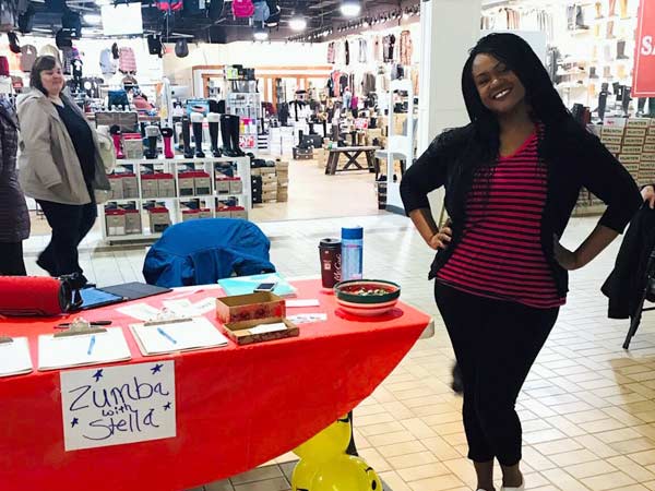 Stella in the mall. Beside her is a table with a sign that says "Zumba with Stella."