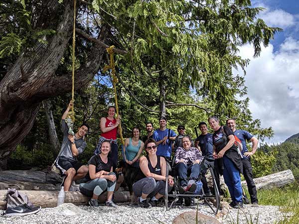 A group of people on a beach at the edge of large trees, with a woman sitting in the Trail Rider equipment.