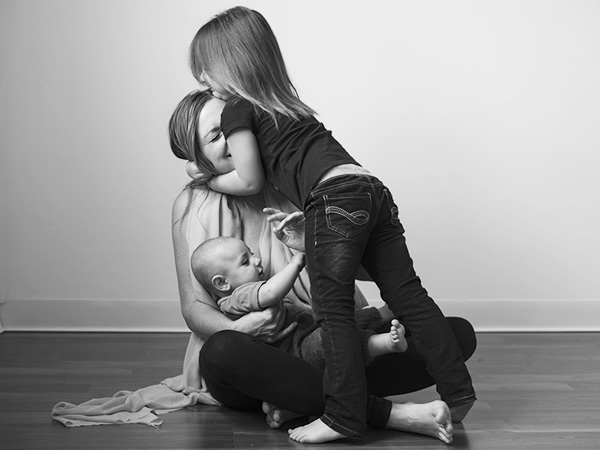Black and white image of woman sitting on the floor, holding her infant son while her daughter hugs her.