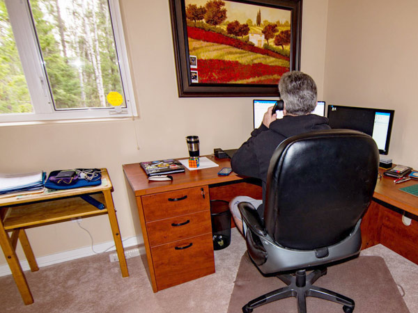 A man faces his desk and two monitors in his home office. To his right, a coffee and Rubiks cube sit under a window. Outside is a forest. 