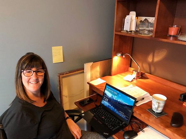 A woman smiles into the camera, her laptop sits on her wood desk at home.