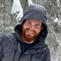 Man with ginger beard standing in the snow with gray jacket and hoodie on.