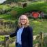 Woman with shoulder length blonde hair and glasses stands in front of hobbit house