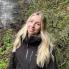 Woman with long blonde hair, wearing a black rain coat in a wooded location with a waterfall in the background.