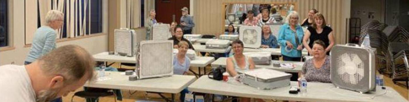 People sit at long tables with DIY air cleaners standing next to them.